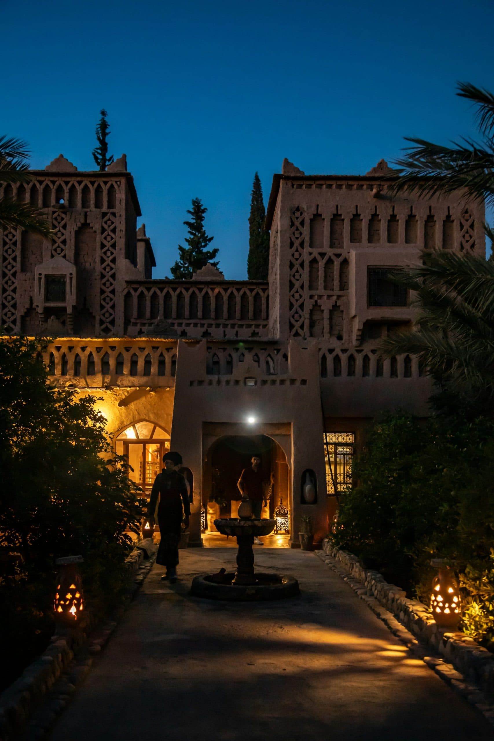 A Moroccan kasbah at blue hour, a lantern-lit path leading to a warm, glowing courtyard entrance