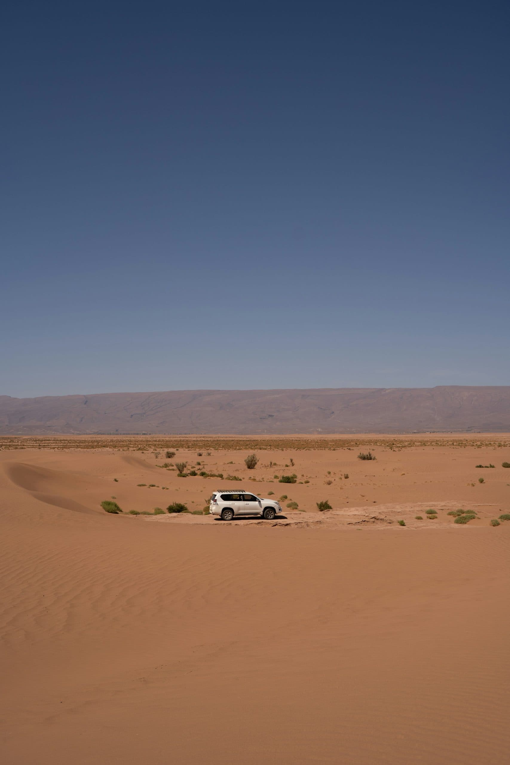 A white 4×4 crossing open Saharan sand below the bare ridgeline of the Draa Valley